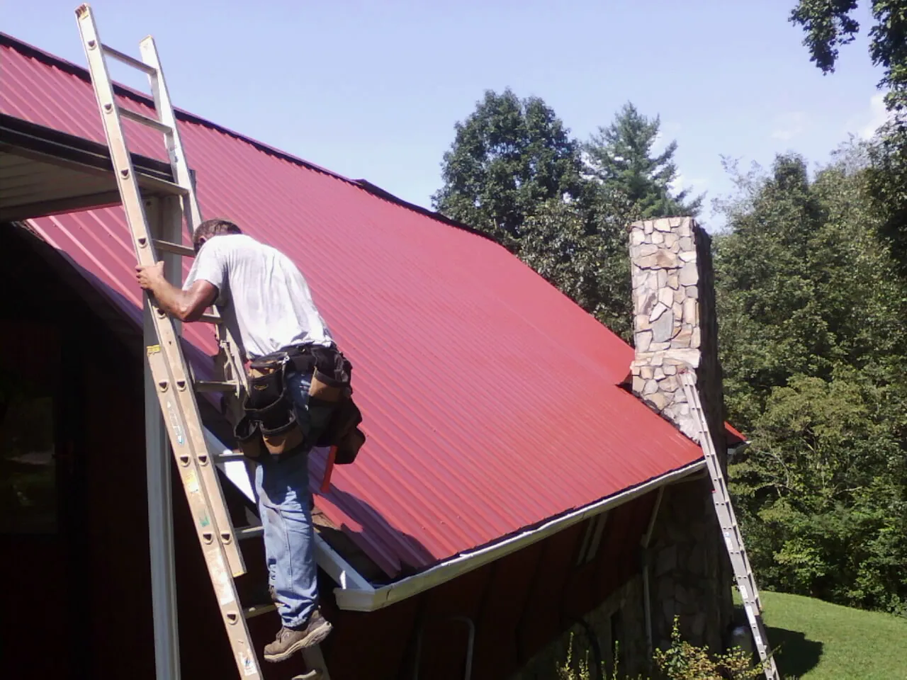 Roofer on a ladder beside a red metal roof and stone chimney — Soffit & Fascia Repair by Roofing Brothers in Deptford